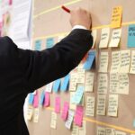 A man in a suit writes on a board covered with sticky notes, illustrating a knowledge management tool in action.