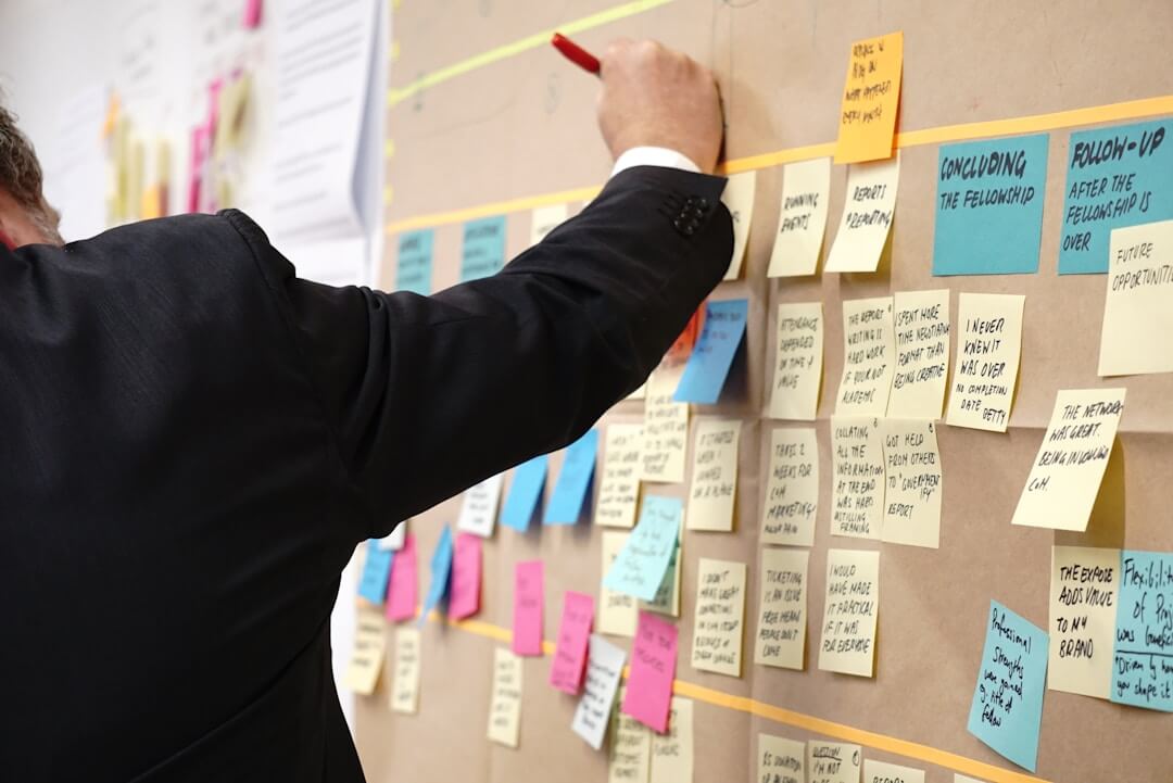 A man in a suit writes on a board covered with sticky notes, illustrating a knowledge management tool in action.